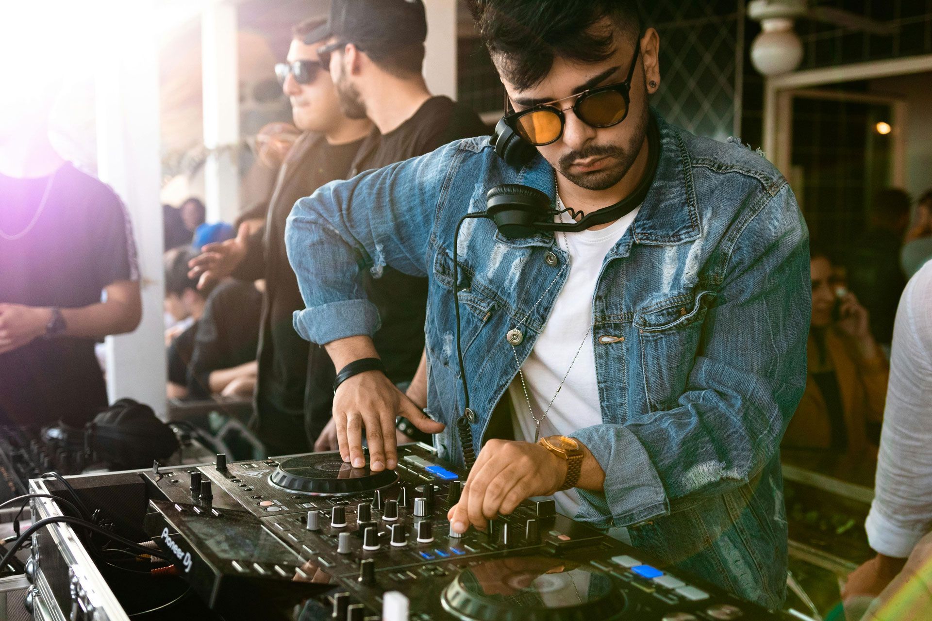 A DJ in a denim jacket adjusting a mixing console while standing at an outdoor event.