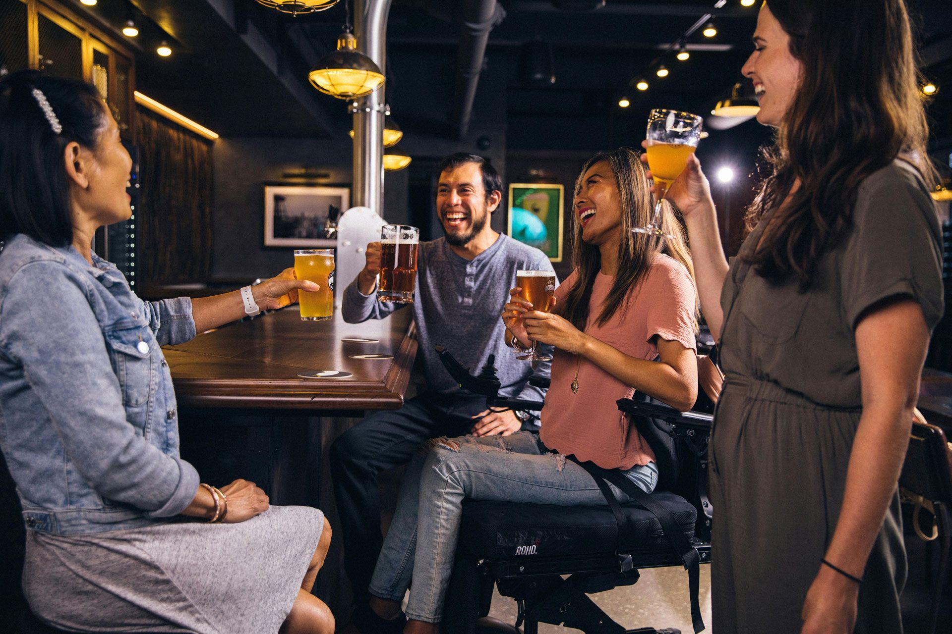 Four friends toast with glasses of beer while laughing together at a bar table.