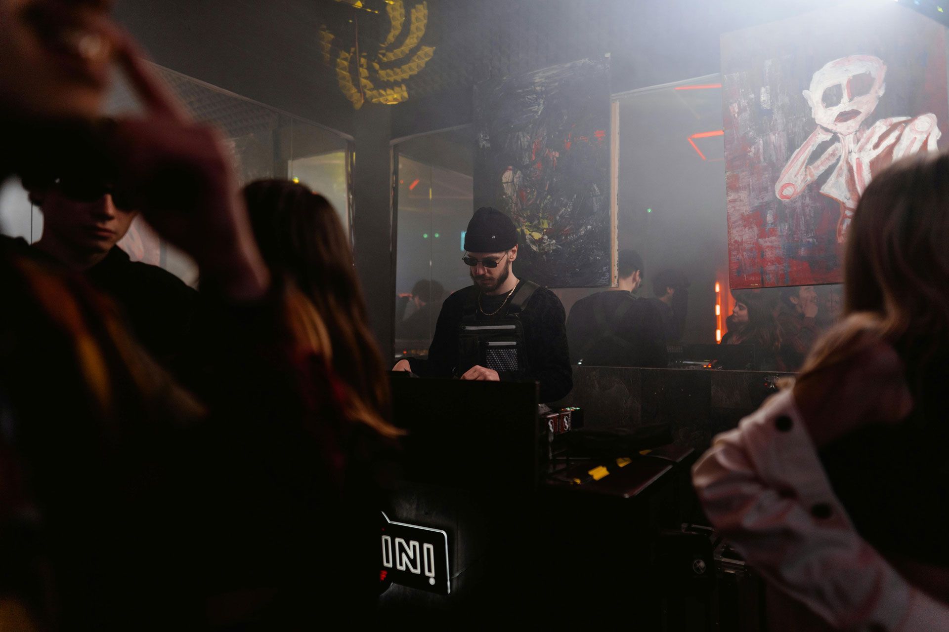 A DJ performs at a dimly lit club, surrounded by people in a hazy, atmospheric room with wall art.