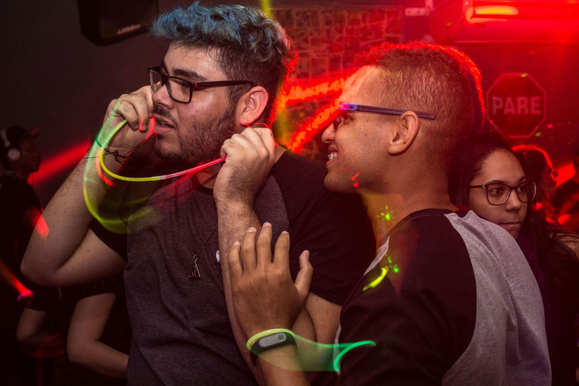 Two people interact at a dimly lit club, one holding a glowing neon accessory near their face while smiling at the other.