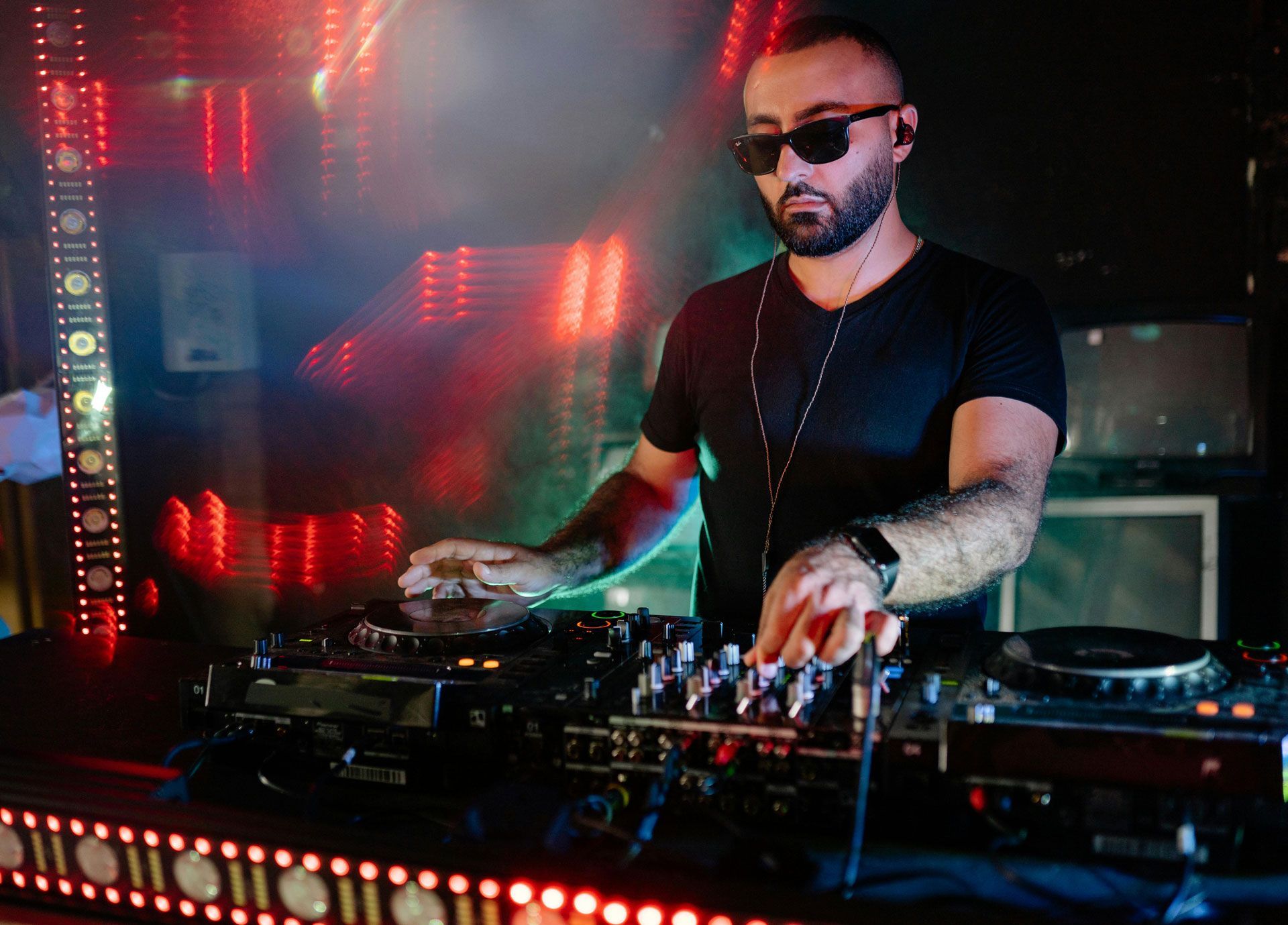A DJ in sunglasses performs on stage behind a mixing console, illuminated by vibrant red and purple nightclub lighting.