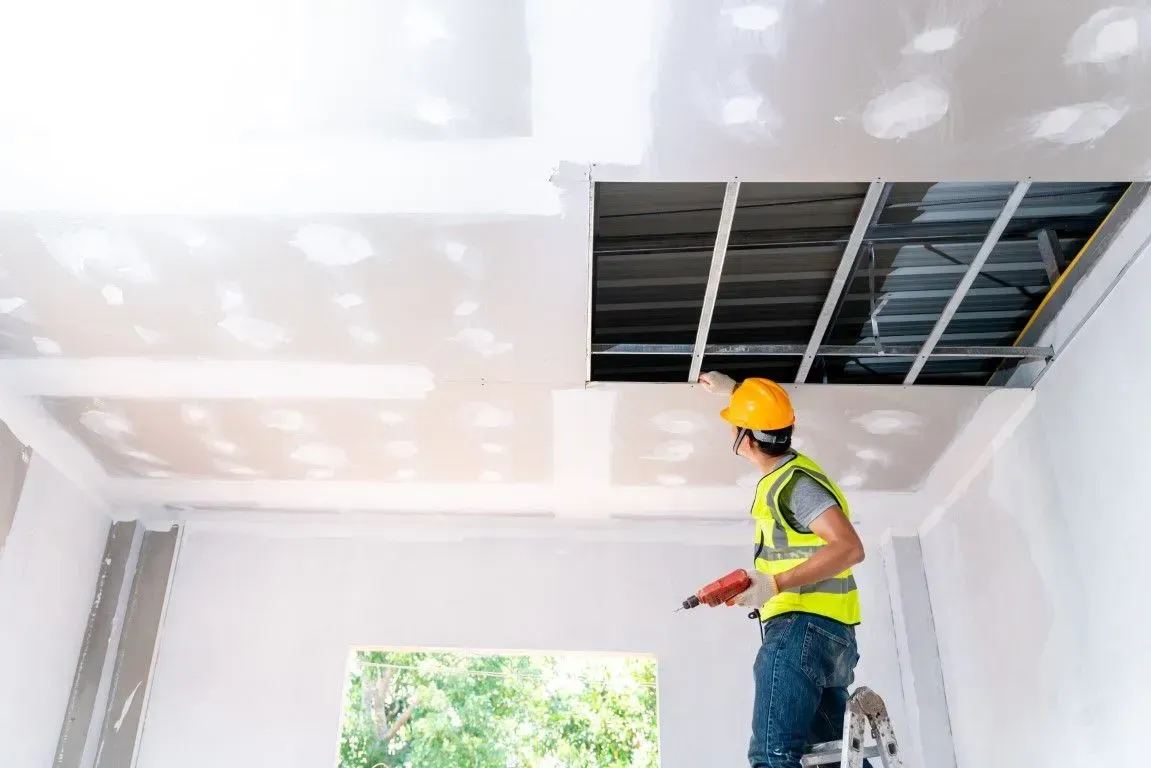 Contractor in hard hat installing drywall panels on a ceiling.