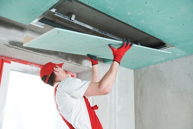 Worker installing a ceiling panel in a room, wearing red gloves and cap.