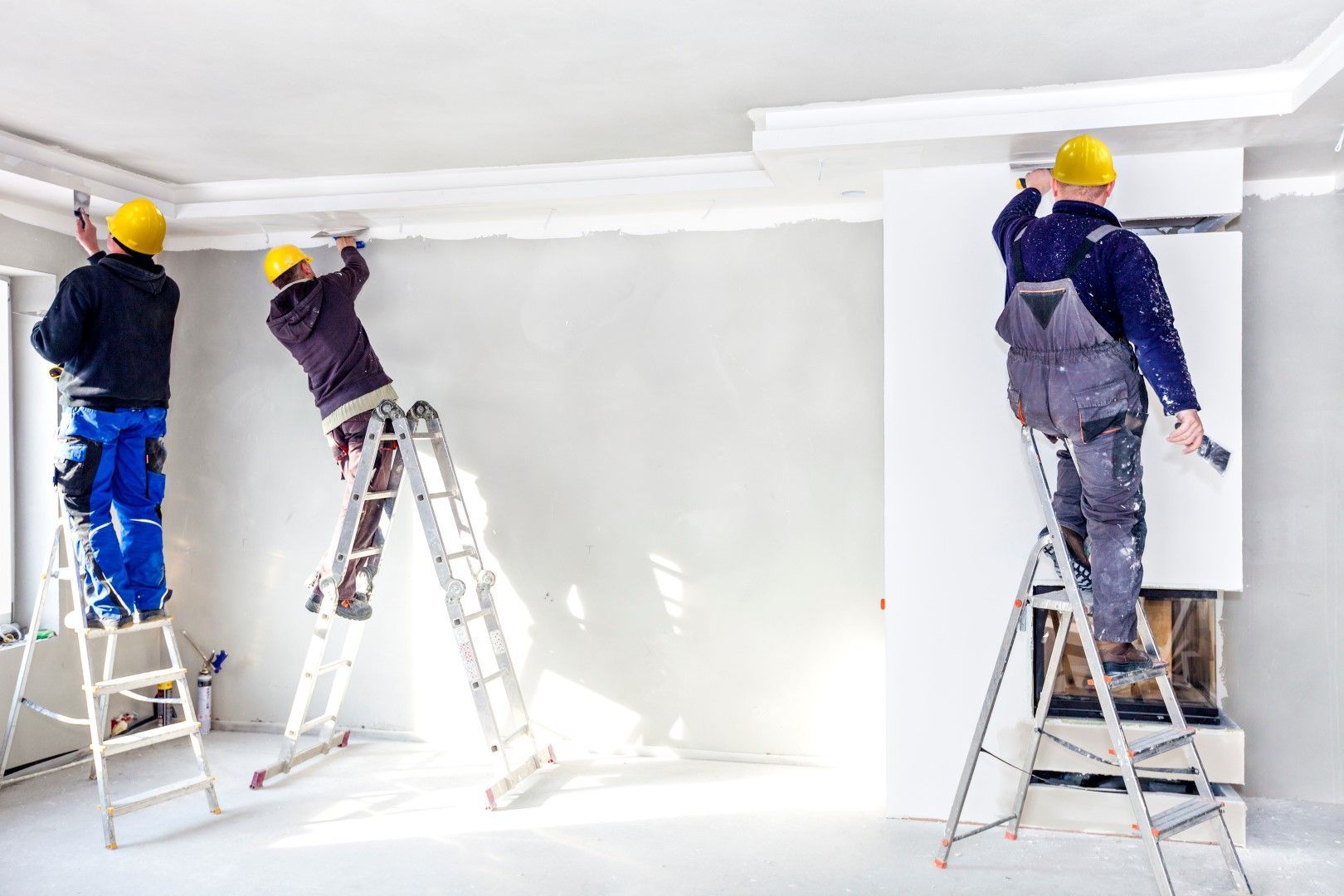 Three workers on ladders installing crown molding on drywall.