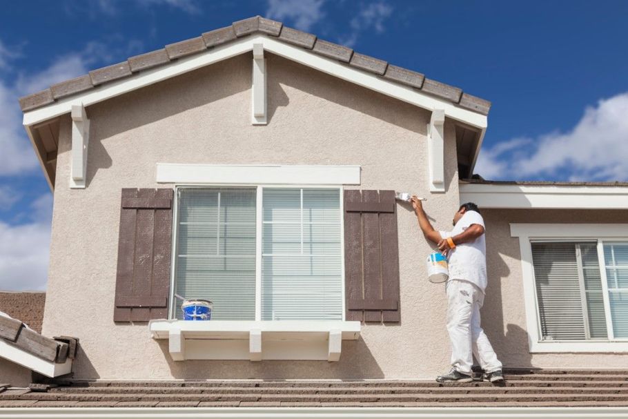 Worker painting the exterior trim of a beige house under a blue sky