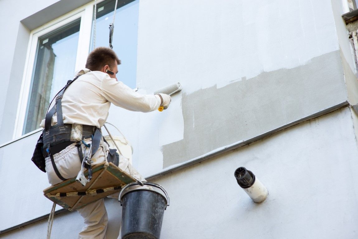 Painter on a scaffold applying light gray paint to an exterior wall beside a window.