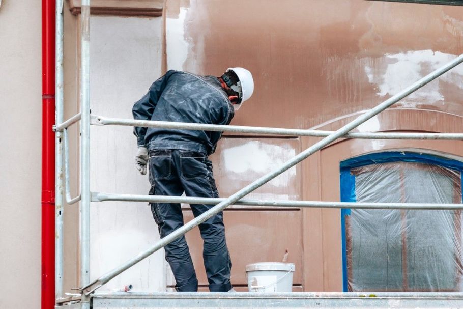 Worker in dark coveralls painting a building facade from scaffolding beside a white bucket.