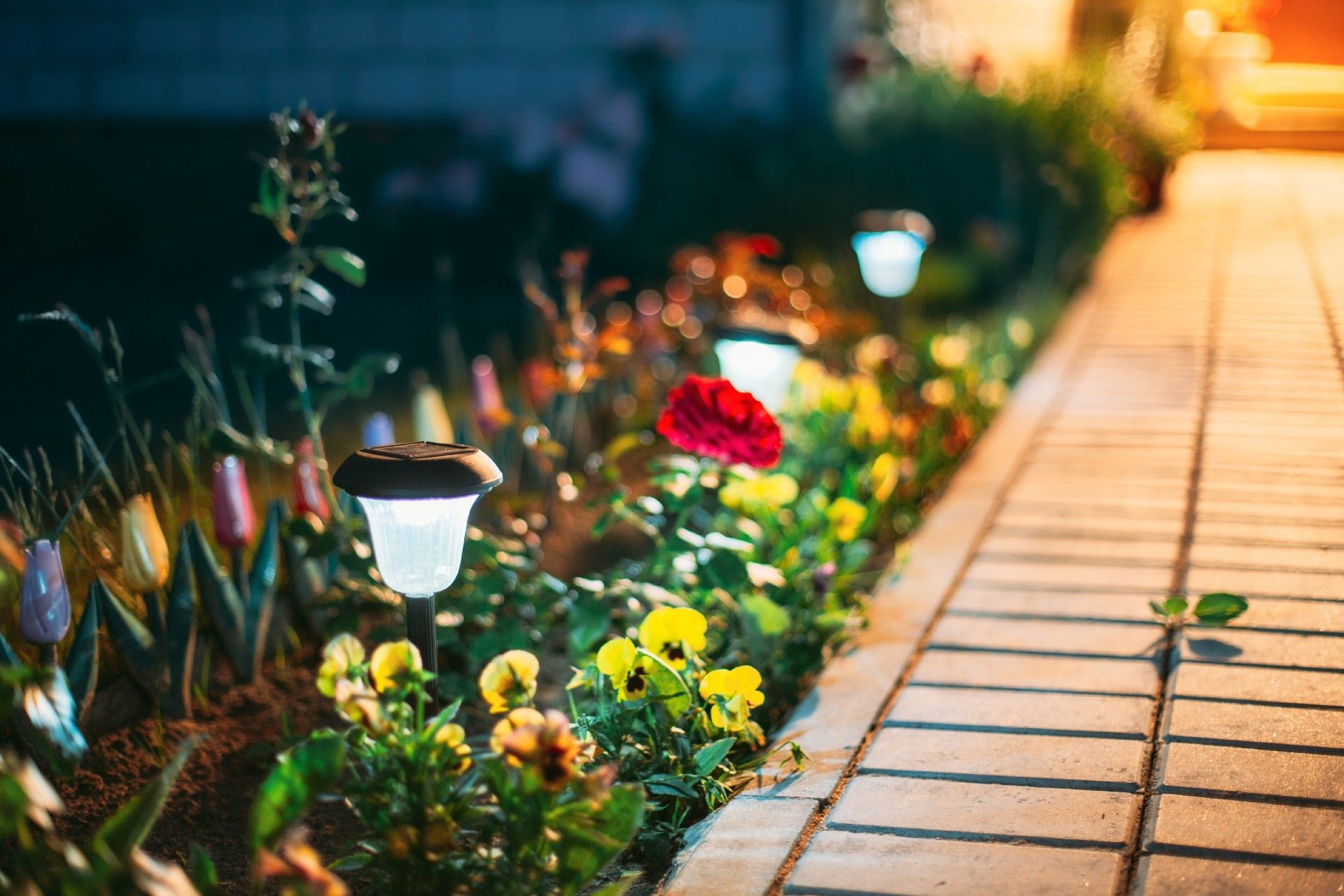 A brick walkway leading to a garden with flowers and solar lights.