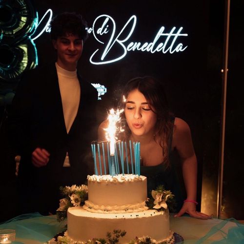 Una donna che spegne le candeline su una torta davanti a un cartello con la scritta benedetta