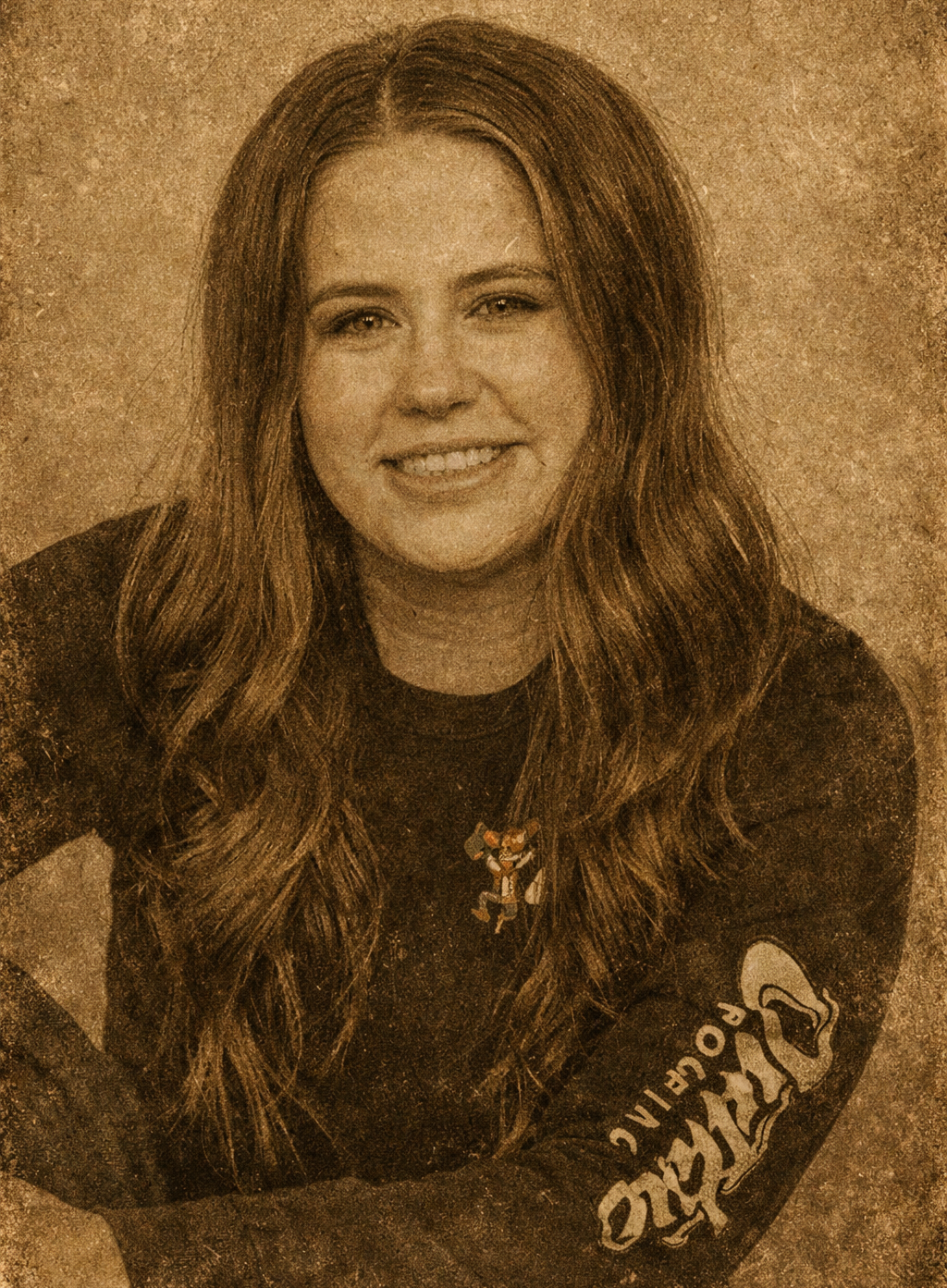 A sepia-toned headshot of a smiling person with long, wavy hair wearing a long-sleeved shirt with branded text on the arm.