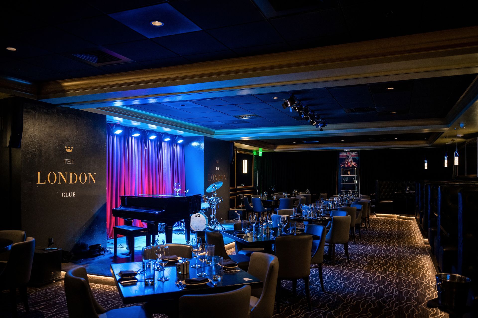 Inside a dimly lit jazz club with a stage. Grand piano, drum set, tables, and chairs. Blue and purple lighting.