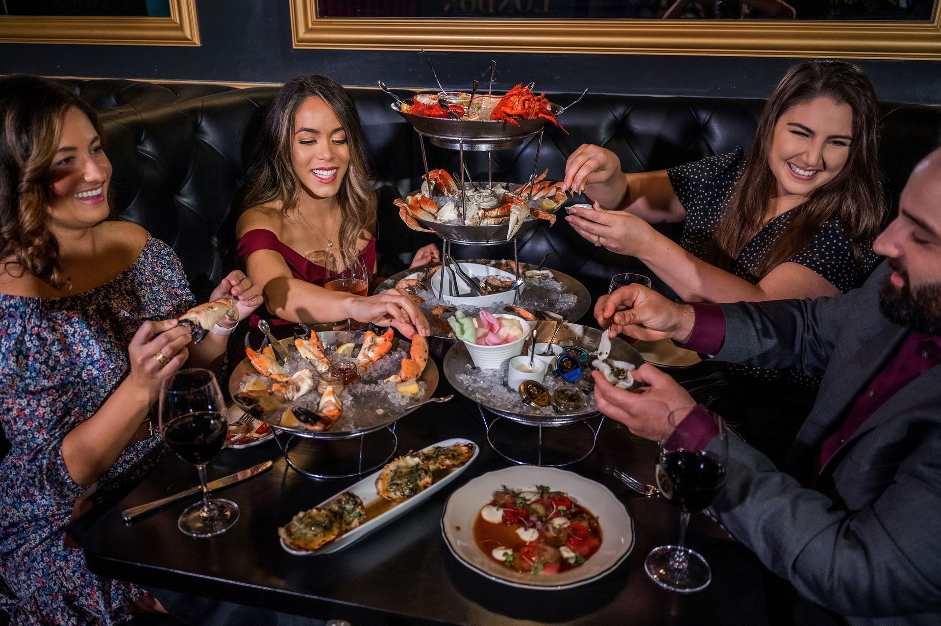 Four people at a table enjoying seafood. They laugh while eating; ornate plates, wine, and a dark setting.