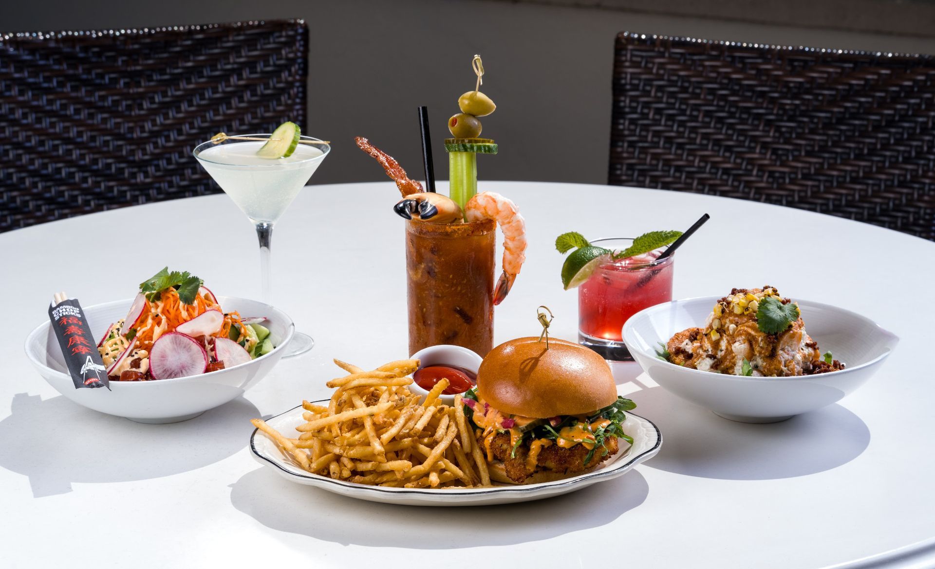 Table with drinks and food, including a burger, fries, salads, and cocktails, in an outdoor setting.