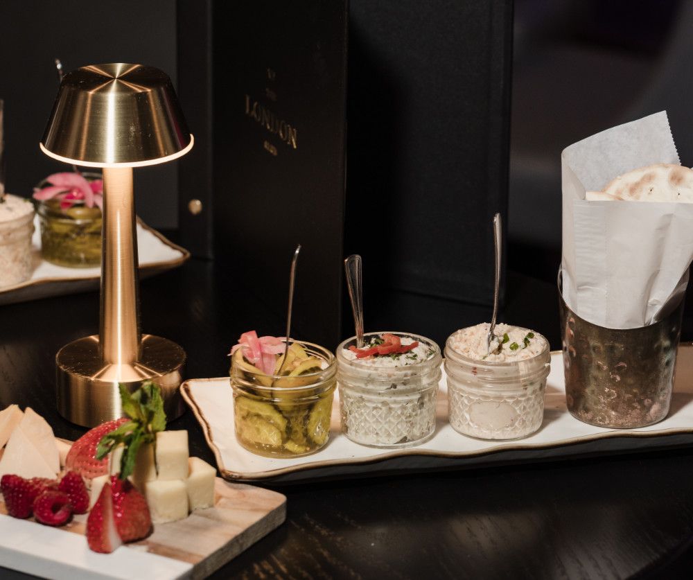 Appetizers with pickles, dip, cheese, and bread served on a tray with a golden lamp in a restaurant setting.