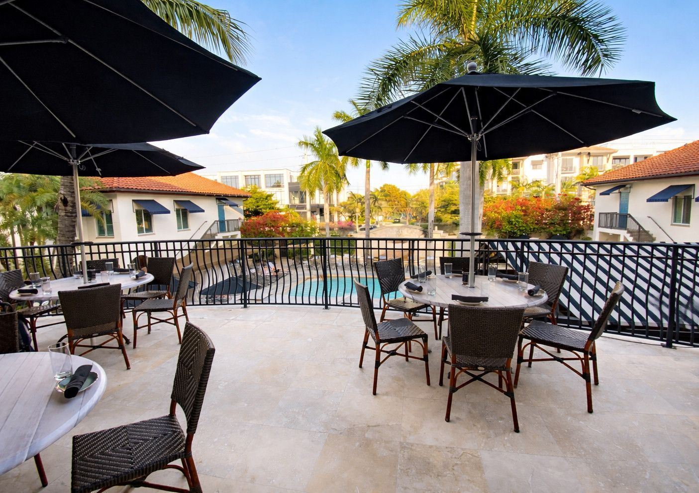 Outdoor patio with tables, chairs, and black umbrellas; a pool and buildings in the background.