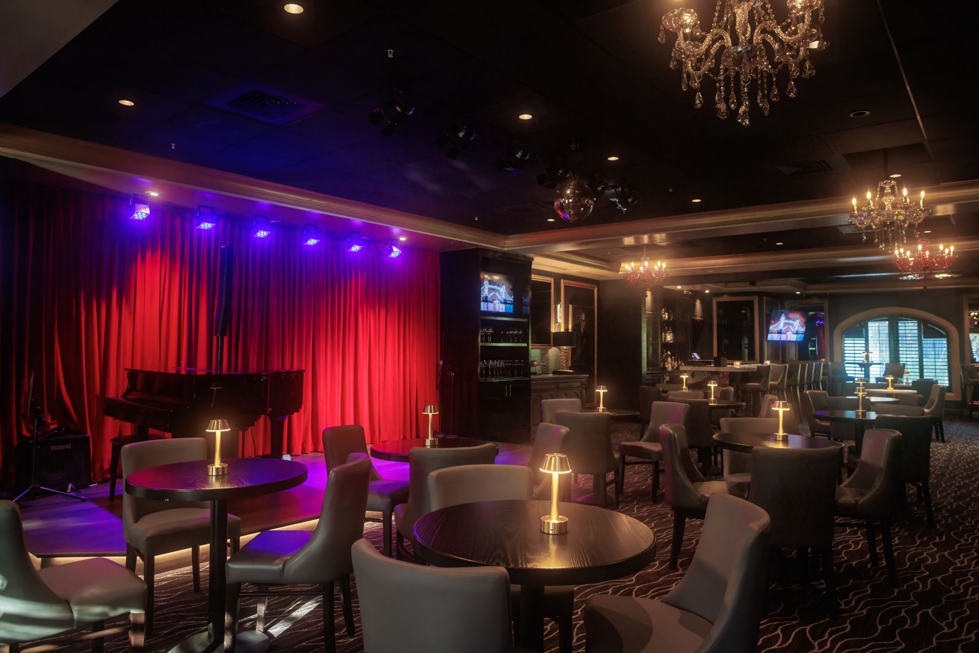 Interior of a dimly lit jazz club with a stage, piano, tables, and chairs. Purple and blue lighting.