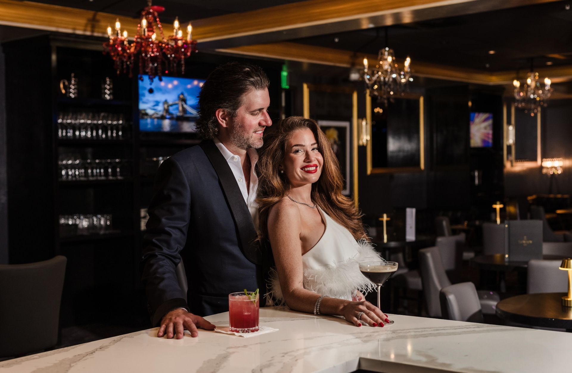 Couple at a bar, man in a suit smiles at woman in white dress, a cocktail rests on the bar.