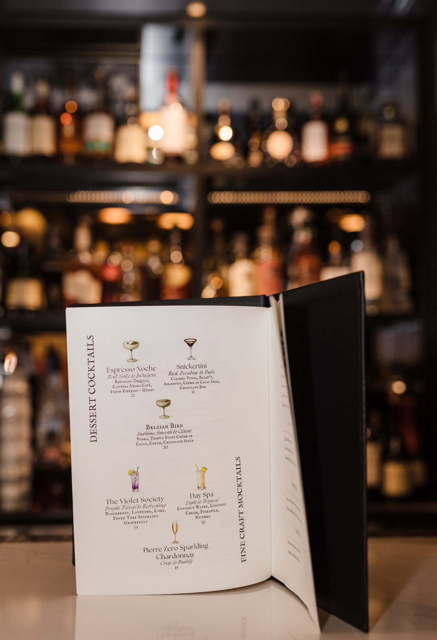 Cocktail menu on a bar, with drinks listed and a blurred liquor shelf in the background.