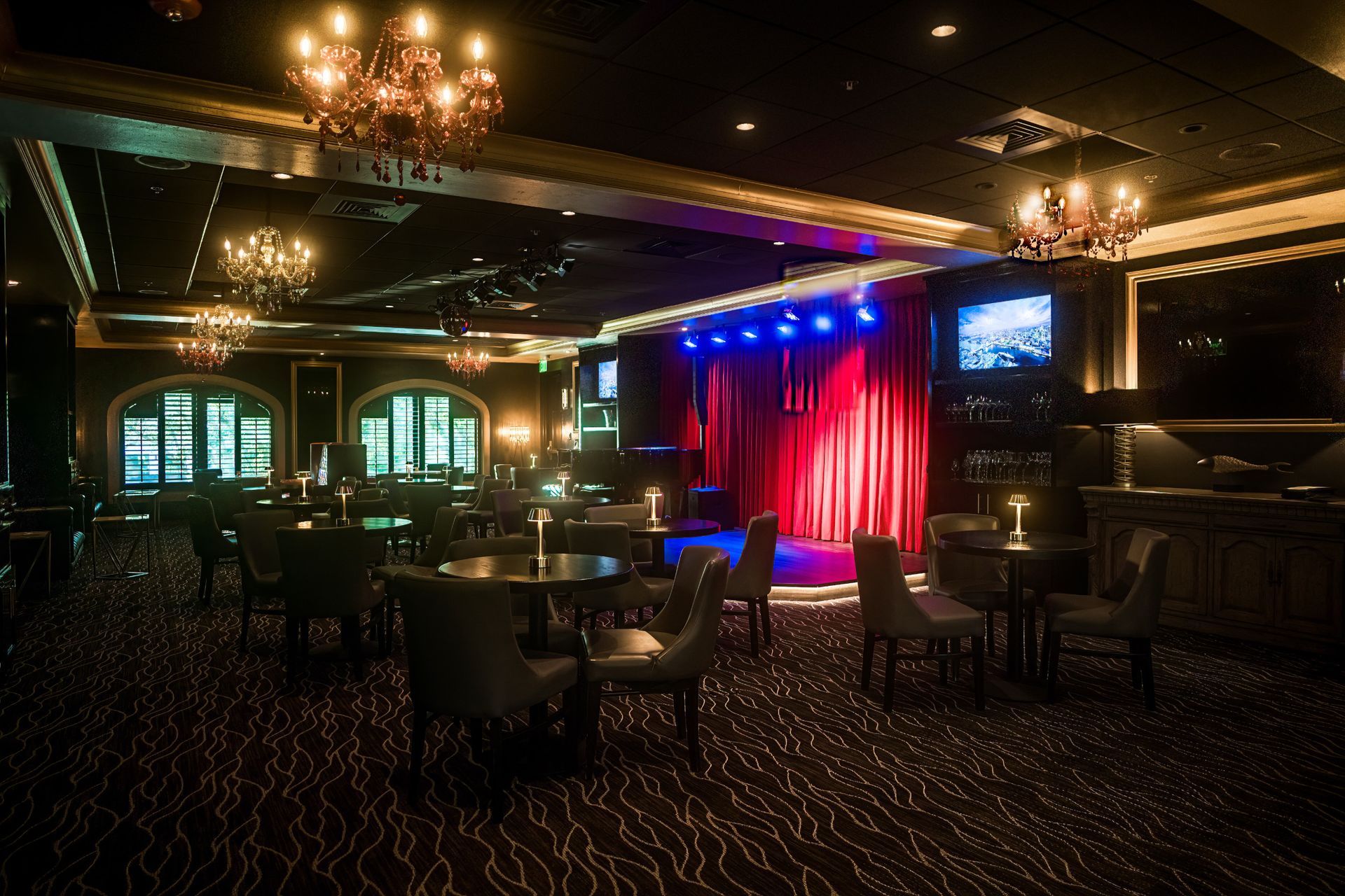 Dark, empty performance space with a red curtain stage, small tables, chairs, and chandeliers.