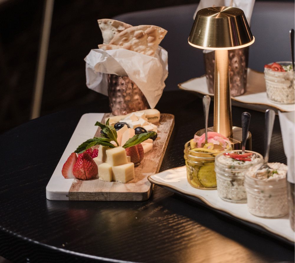 Appetizers on a dark table: cheese board with fruit, crackers, dips in jars, and a lit gold lamp.