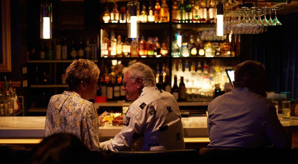 People at a bar, illuminated by soft lighting. Liquor bottles and glasses on shelves behind the bar.