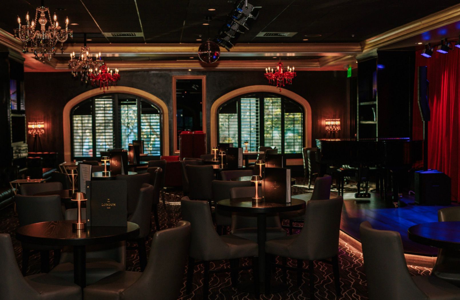 Elegant dimly lit bar with tables, chairs, arched windows, stage with piano, and red chandeliers.