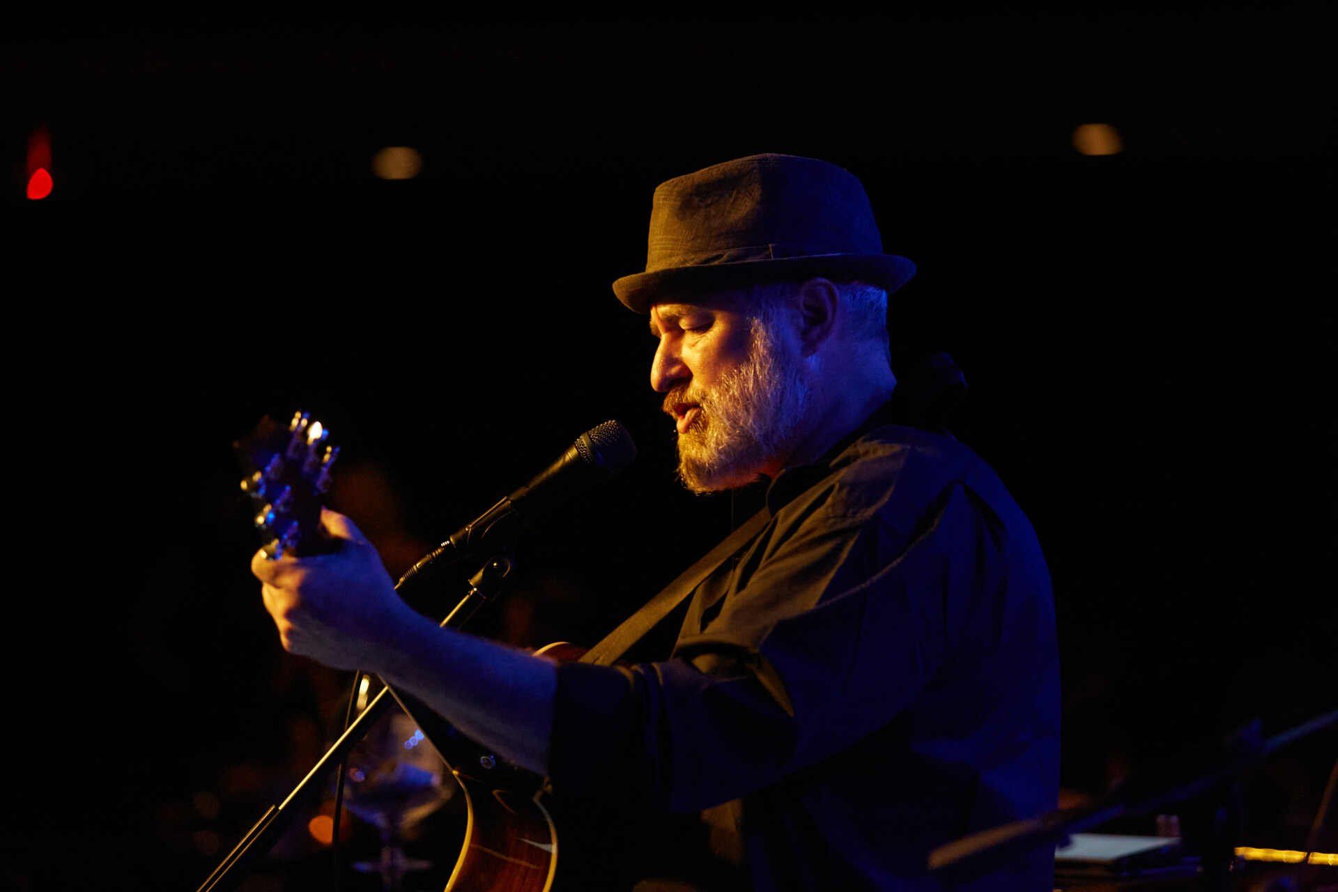 Man with a grey beard wearing a hat, singing and playing a guitar on stage with a dark background.
