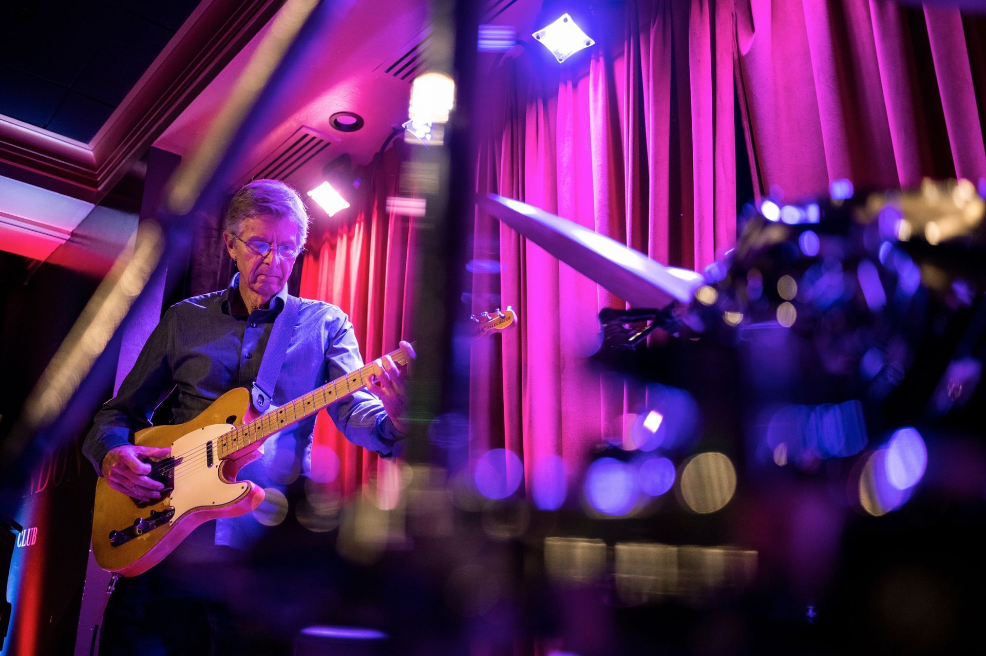 Guitarist playing on stage in front of red curtains; purple and blue stage lighting.