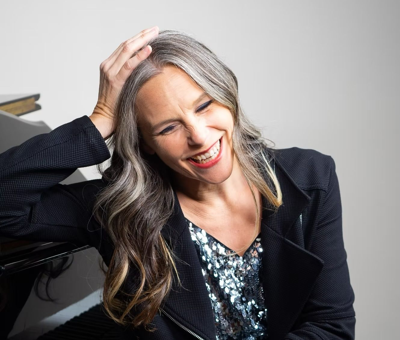 Woman with graying hair laughs, resting on a piano. She wears a sequined top and black jacket.