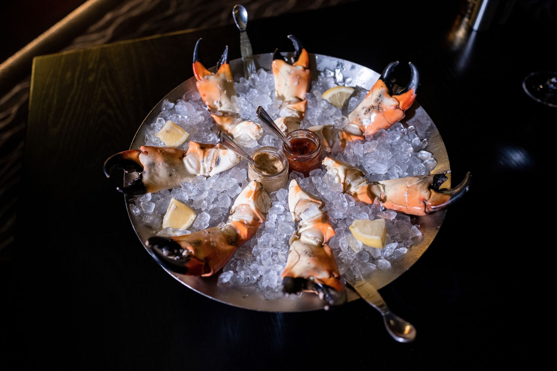 Crab claws on ice with dipping sauces and lemon wedges on a black table.