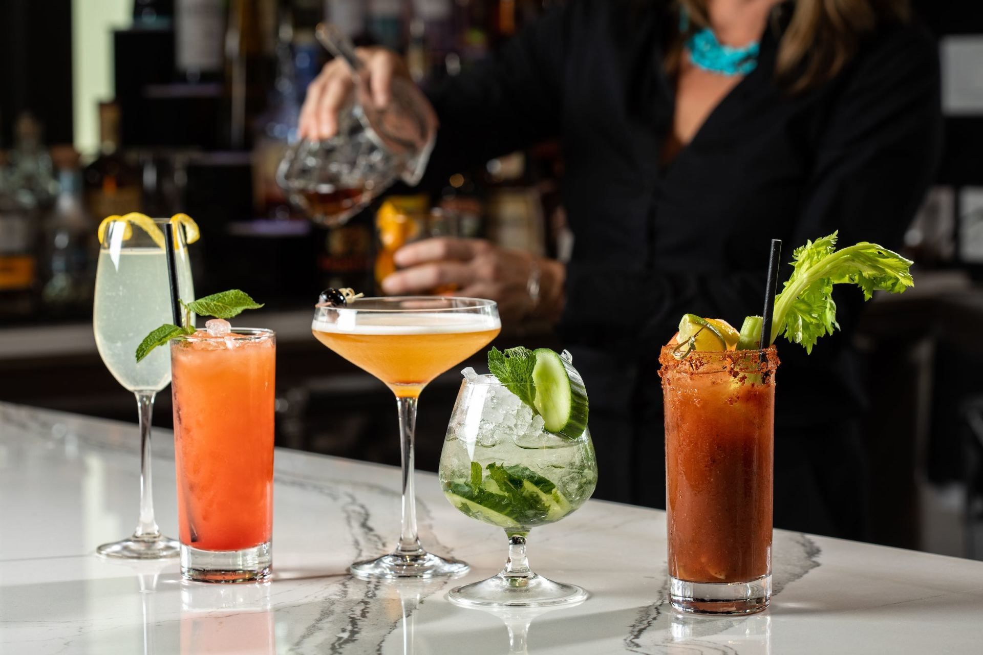 A bartender pours a drink at a bar with five colorful cocktails lined up on the counter.