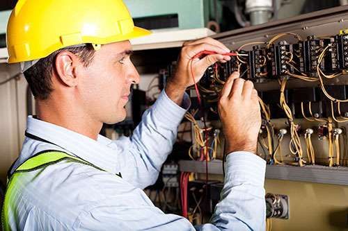 Electrician in a yellow hard hat wiring an electrical control panel with tools