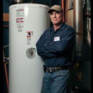 Technician standing beside a white water heater in a utility room, arms crossed.