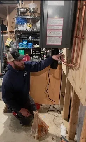 Technician kneeling beside a wall-mounted furnace, inspecting copper pipes in a basement utility room.