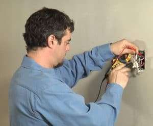 Man working on exposed wall wiring with tools, wearing a blue shirt