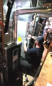 Worker in dark clothing kneels inside an industrial machine, operating controls amid metal pipes and panels.