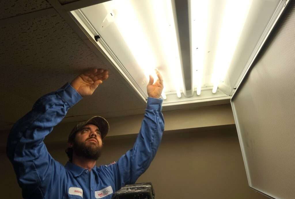 Technician in blue uniform reaching up to replace fluorescent lights in a ceiling panel