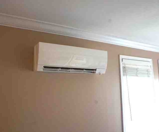 Wall-mounted white air conditioner above a window in a beige room.