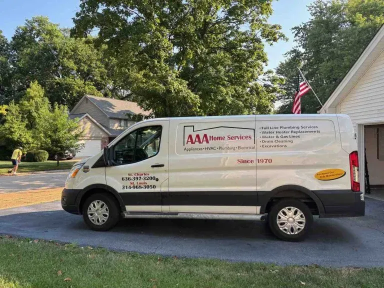 White AAA service van parked in a driveway beside a house, with trees and an American flag in the background
