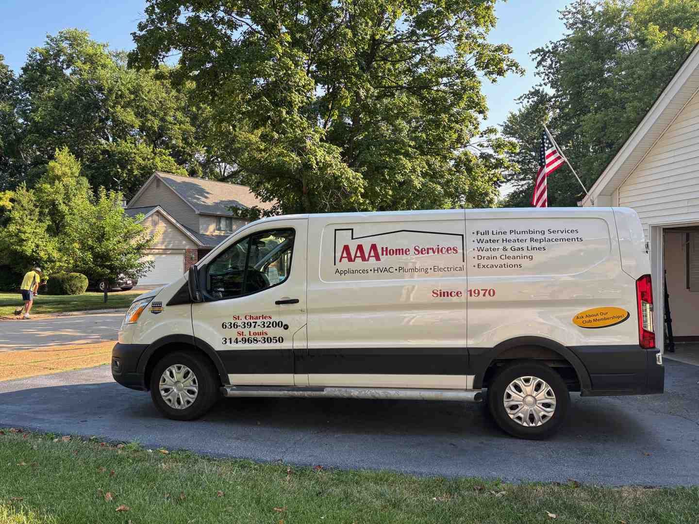 White service van parked in a driveway beside a house, with trees and an American flag in the background