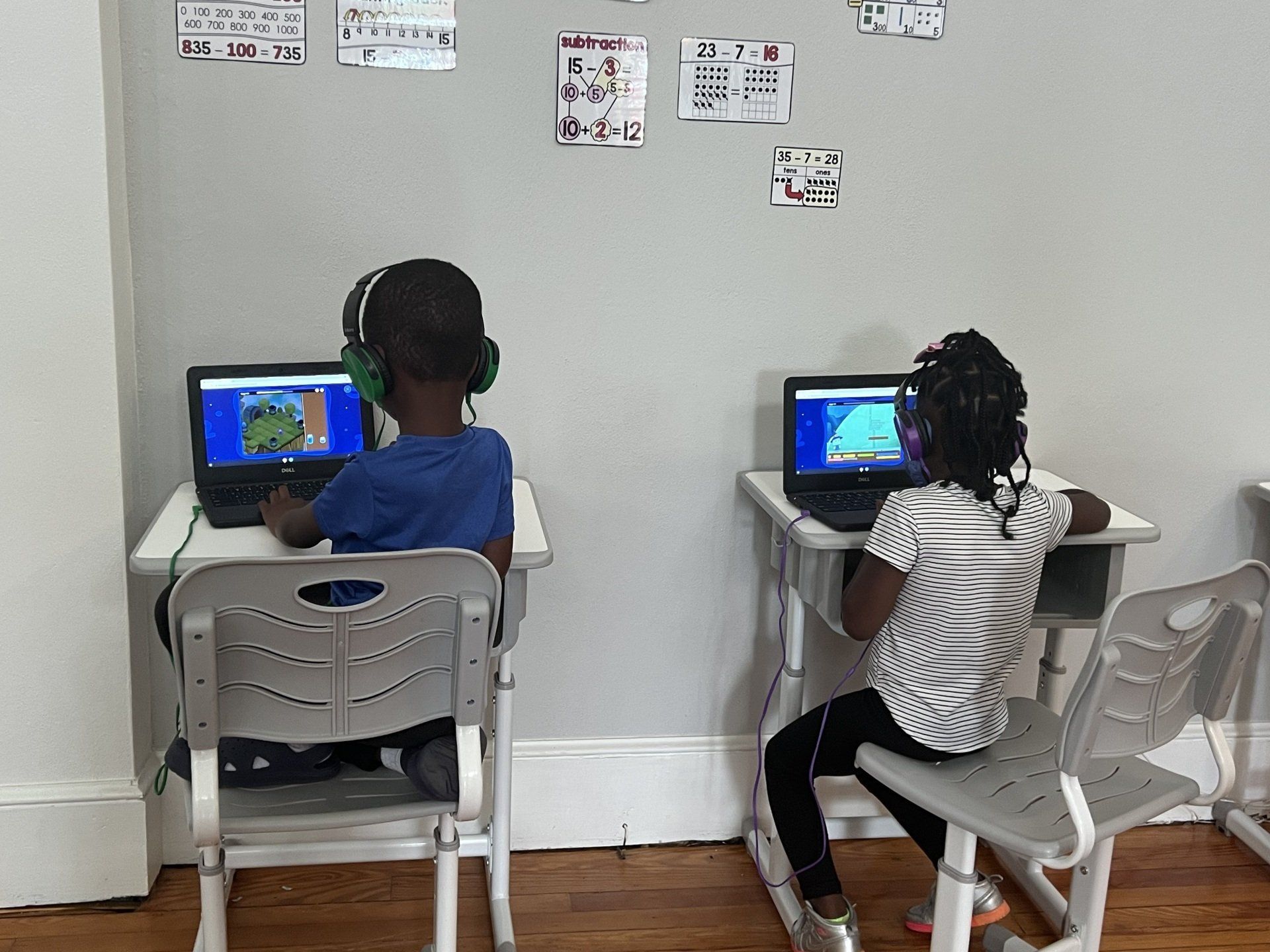 Two children are sitting at desks with laptops in a classroom.
