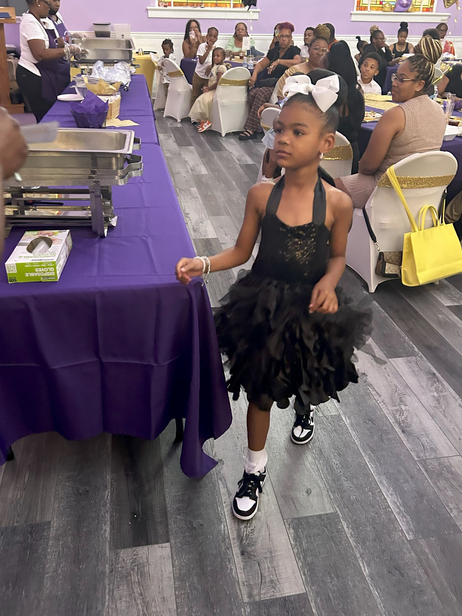 A little girl in a black dress is walking in a room at a party.