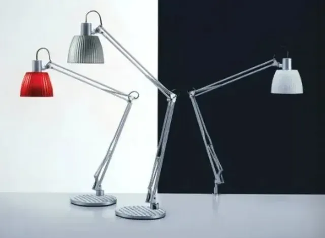 Tre lampade da scrivania con bracci articolati in metallo e paralumi nei colori rosso, grigio metallizzato e bianco, su uno sfondo bicolore bianco e nero.