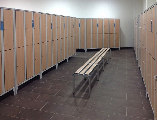 Spogliatoio vuoto caratterizzato da file di armadietti alti in legno chiaro lungo le pareti e una panca centrale in legno su un pavimento piastrellato.