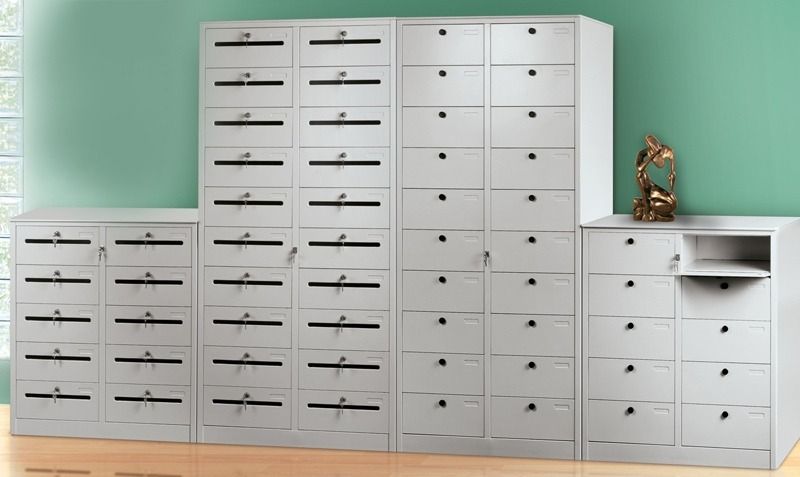 Tre armadietti in metallo grigio con numerosi piccoli scomparti chiudibili a chiave, disposti contro una parete verde.