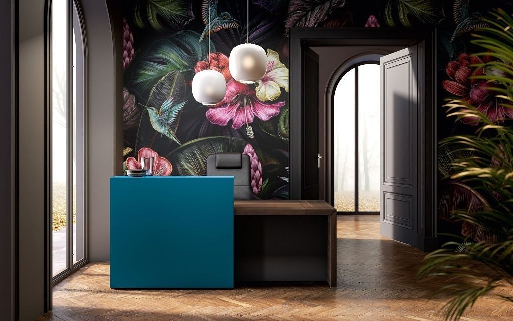 Un bancone della reception color turchese si erge di fronte a una carta da parati scura a motivi floreali, illuminato da due moderne lampade a sospensione e da una porta.