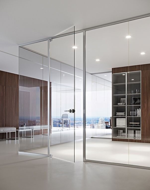 Interno di un ufficio moderno con pareti divisorie in vetro a tutta altezza, una porta in vetro con maniglia scura e una libreria con pannelli in legno.