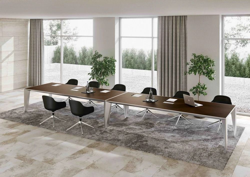 Un lungo tavolo da conferenza in legno scuro con sedie nere, posizionato su un tappeto grigio in un ufficio luminoso e moderno con ampie finestre.