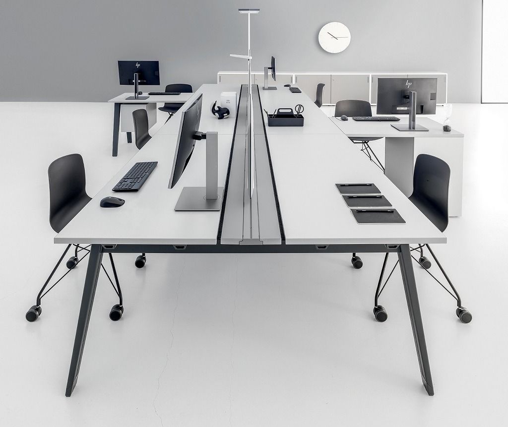 Una scrivania da ufficio bianca a doppia faccia con sedie nere e una lampada divisoria centrale, inserita in una stanza minimalista con un orologio a parete.