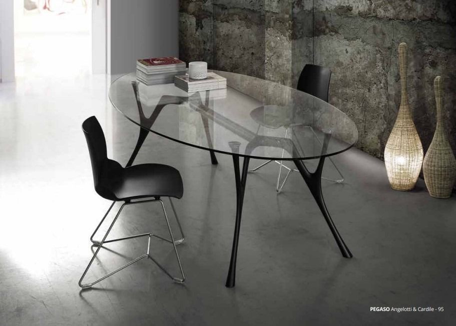 Un tavolo da pranzo ovale in vetro con gambe scultoree nere e sedie moderne nere, collocato in una stanza con una parete in cemento.